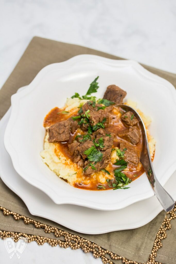 Beef goulash served over mashed potatoes in a white bowl on light brown mat