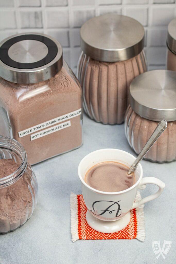 Canisters of hot chocolate mix with white cup of hot chocolate with spoon in it.