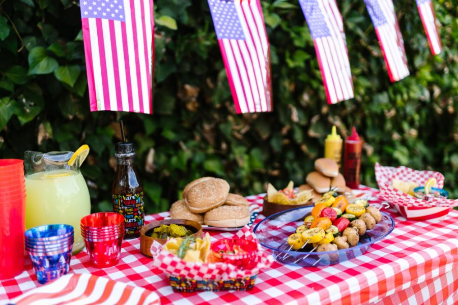 Festive 4th of July picnic table on red checkered tablecloth including kebabs, hamburgers