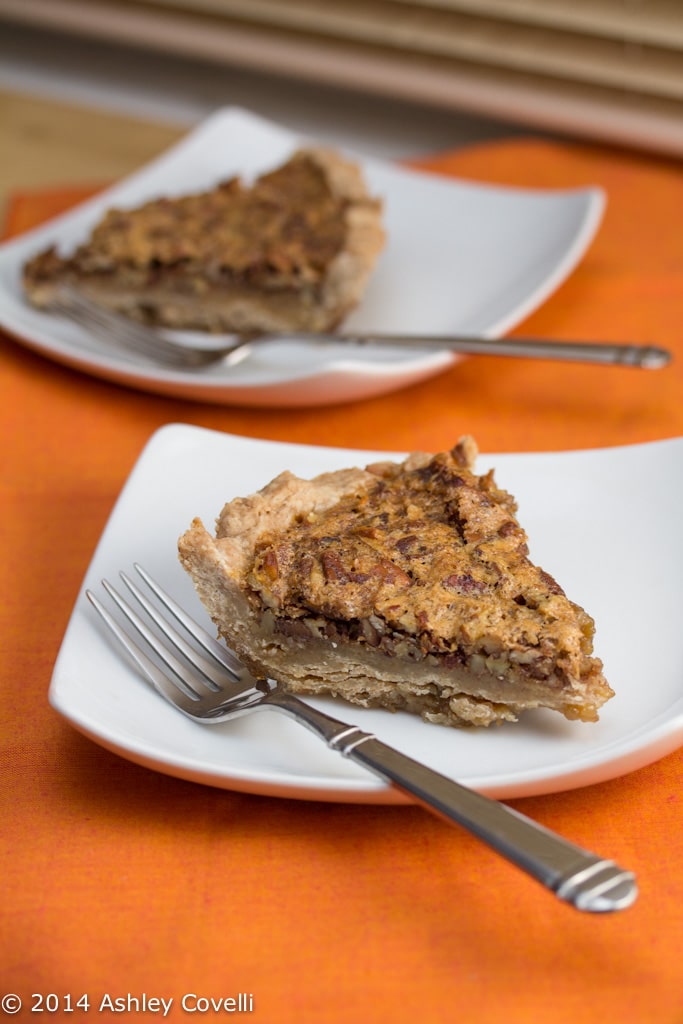 Piece of pecan pie on white plate with fork. On top of an orange tablecloth.